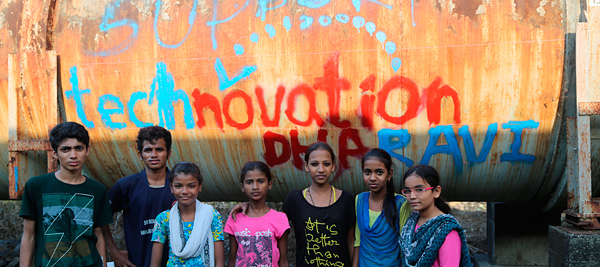 Iridescent's Technovation program logo painted by hand on rusty storage tank in India, with girls in front